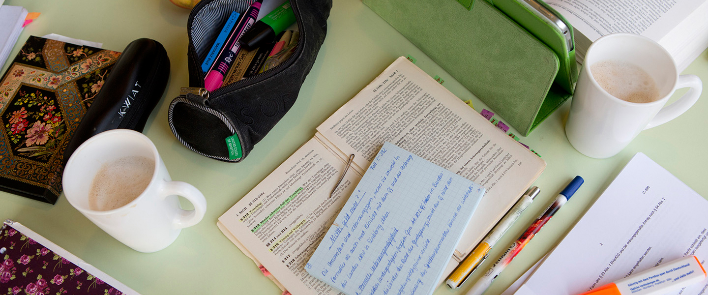 Auf einem Tisch liegen ein Gesetzestext und eine selbst geschriebene Karteikarte, daneben zwei Kaffeetassen und einige Stifte und Blätter, sowie ein Mäppchen, ein Tablet und ein Notizbuch. 